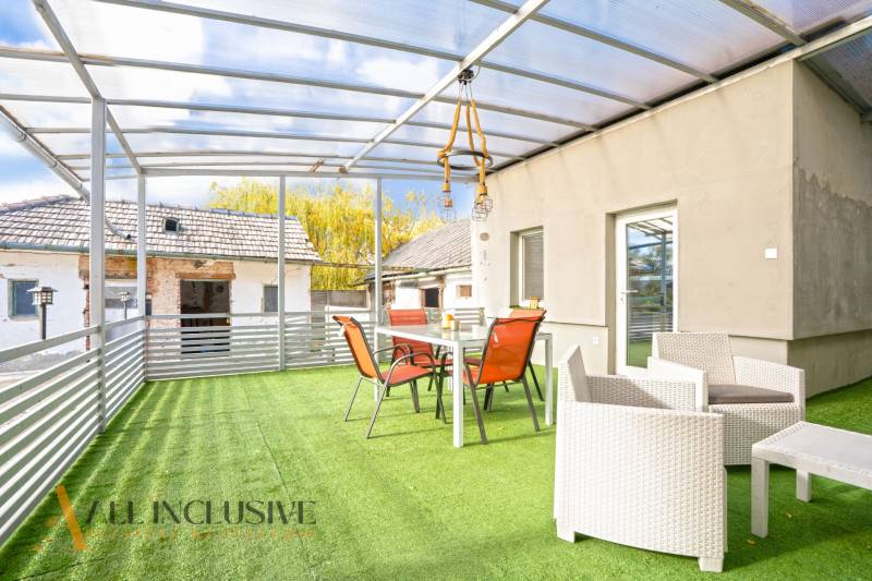 A Nagykeszin található családi ház terasza üvegtetővel és műfüvön elhelyezett bútorokkal.