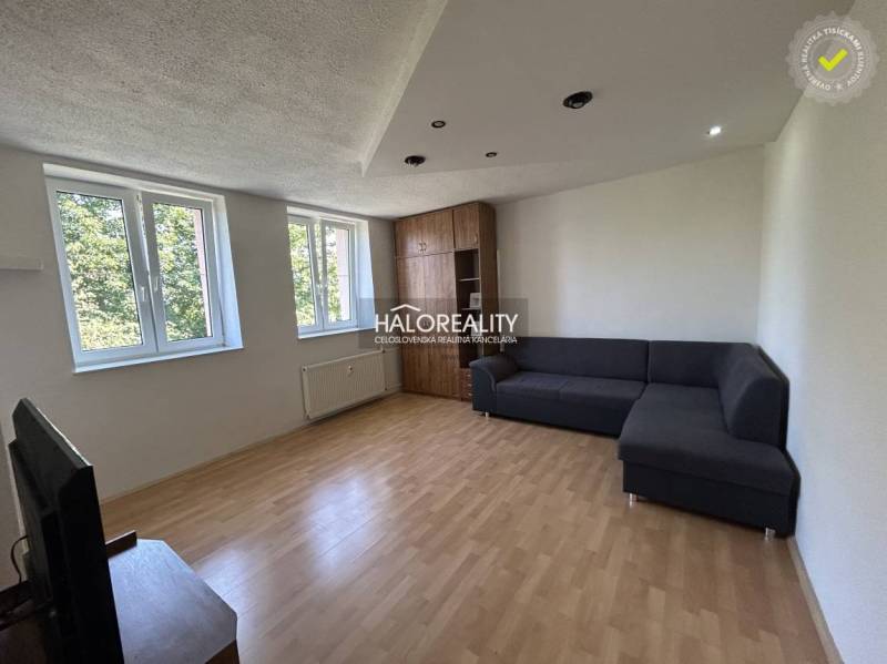 Nappali sötét kanapéval, fa padlóval és két ablakkal egy egyszobás lakásban.