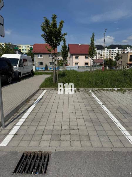 Parkolóhely Kysucké Nové Mesto-ban, háttérben lakóházak és zöldterület.