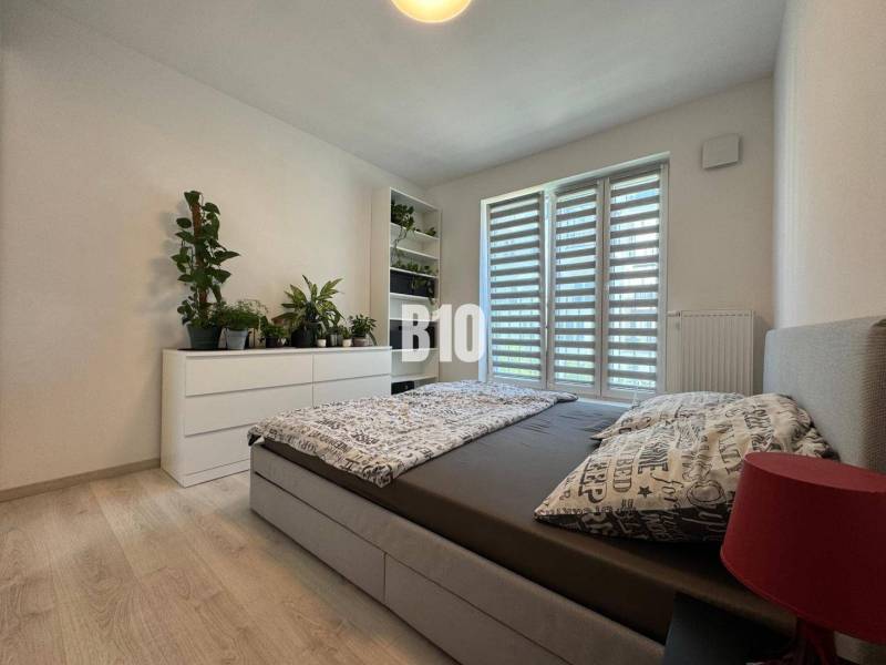 Világos hálószoba fa dekor padlóval, növényekkel, ággyal és fehér komóddal egy kétszobás lakásban.