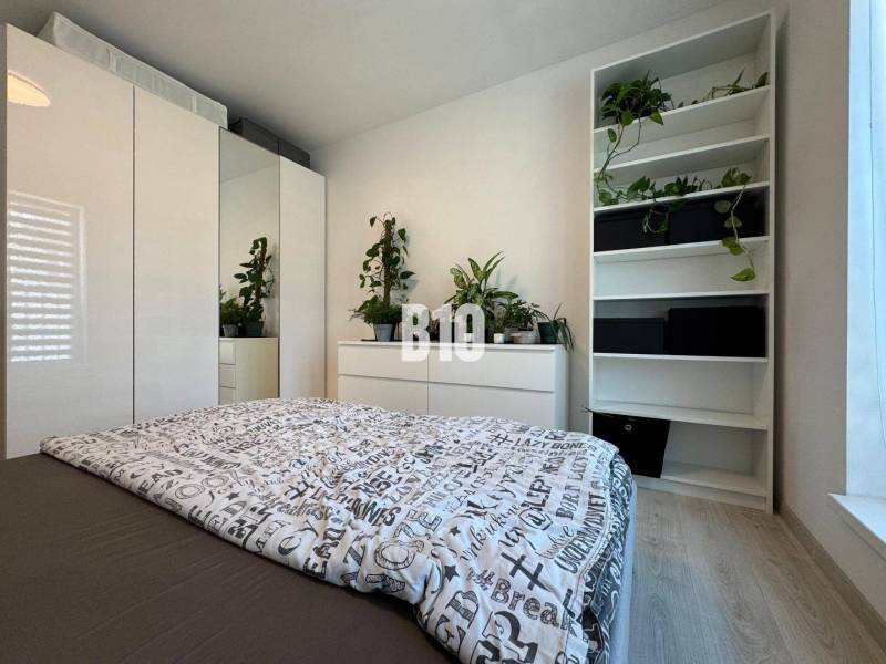 Világos hálószoba egy kétszobás lakásban, nagy szekrénnyel és polcokon lévő növényekkel.