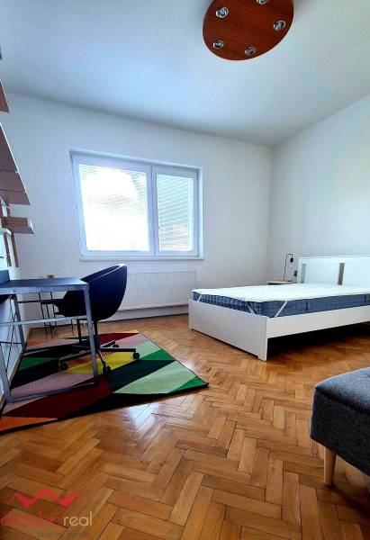 Szoba egy háromszobás lakásban, ággyal, íróasztallal és fa mintázatú padlóval.