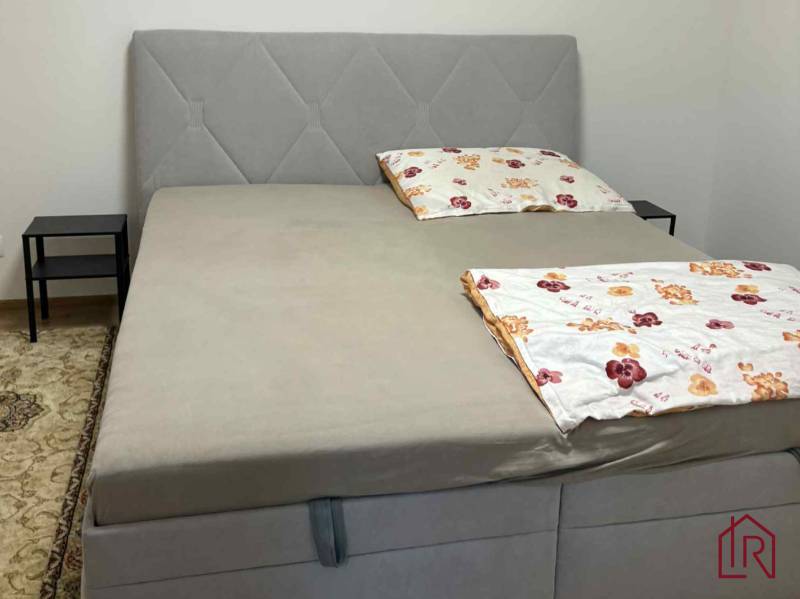 Szürke ágy kárpitozott fejvéggel egy kétszobás lakásban, éjjeliszekrénnyel és szőnyeggel.