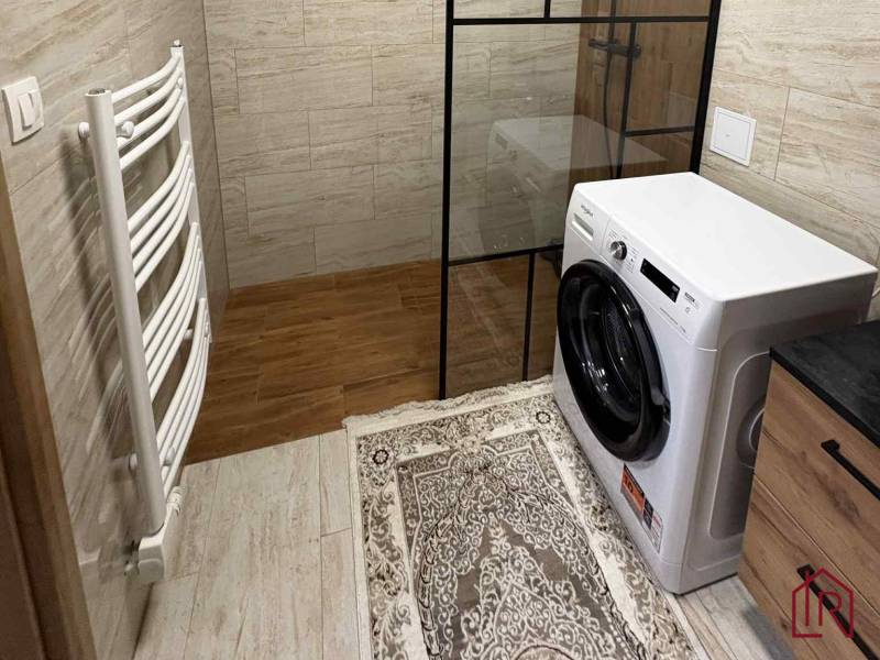 Fürdőszoba egy kétszobás lakásban mosógéppel, zuhanyzóval és fa mintázatú padlóval.