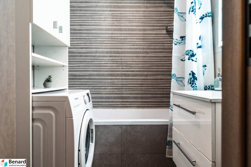 Fürdőszoba egy kétszobás lakásban zuhanyfüggönnyel, mosógéppel és tárolóhellyel.