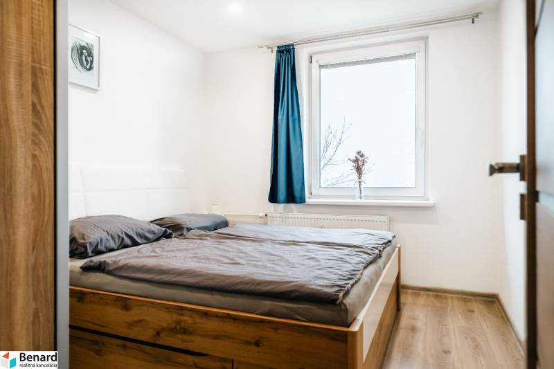 Hálószoba fa mintás padlóval és ággyal egy kétszobás lakásban, kék függöny az ablakon.