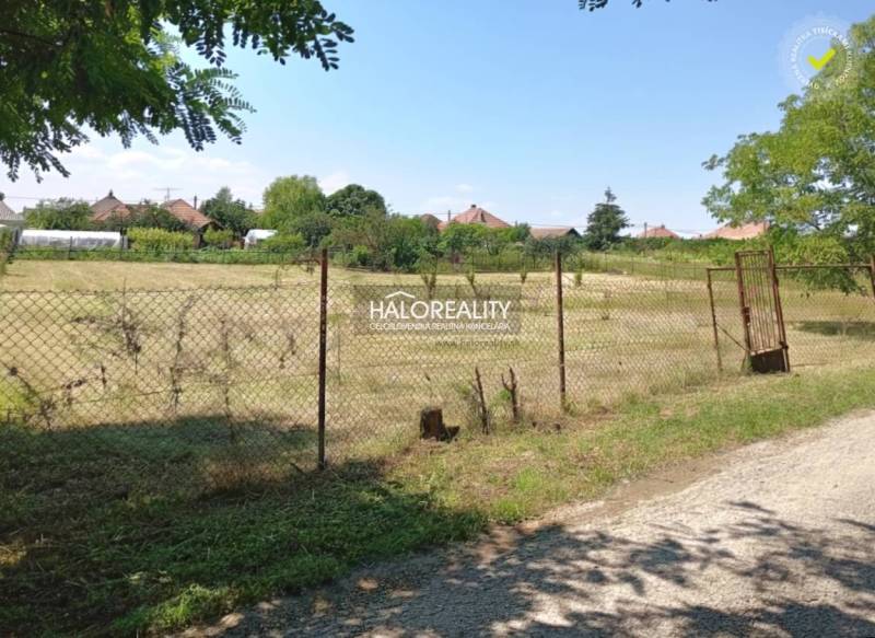 Sík telek kerítéssel körülvéve Marcelová Kertekben, a háttérben házak láthatók.