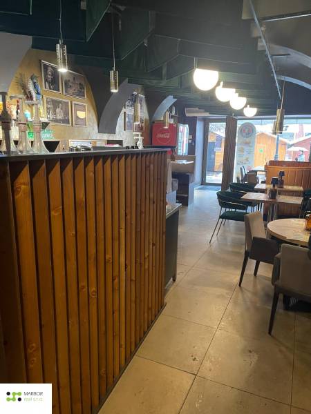 Vendéglátóhely belső tere fa bárral és asztalokkal. Dekoratív világítás is látható.