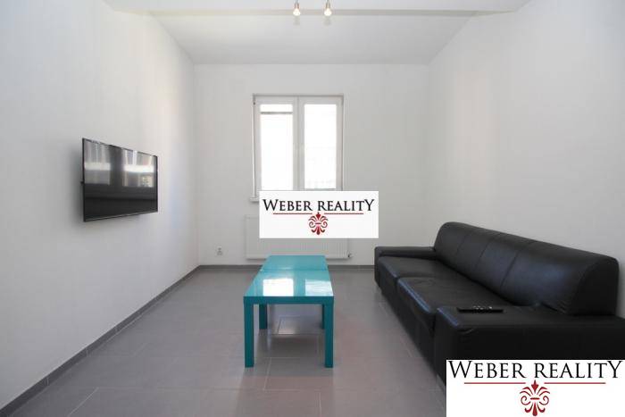 Nappali egy kétszobás lakásban fekete kanapéval, falra szerelt televízióval és alacsony asztallal.