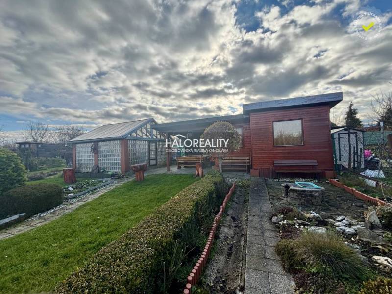 Fából készült házikó a kertekben Iglón, gondozott gyep és bokrok veszik körül.