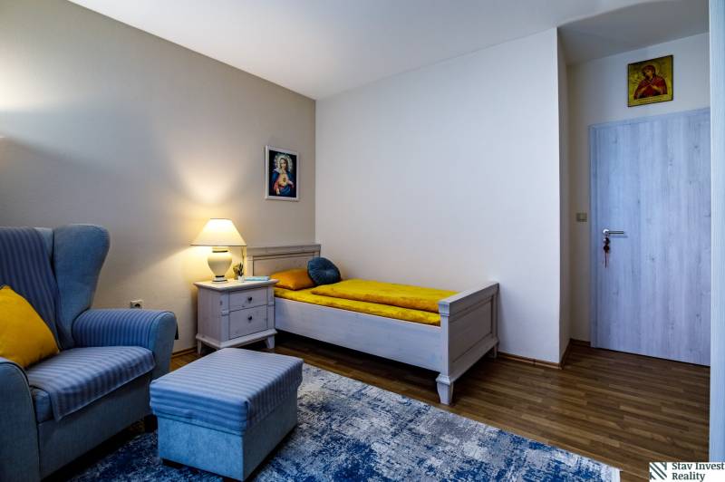 Családi ház szobája fotellel, ággyal, éjjeliszekrénnyel és fa dekorációjú padlóval.