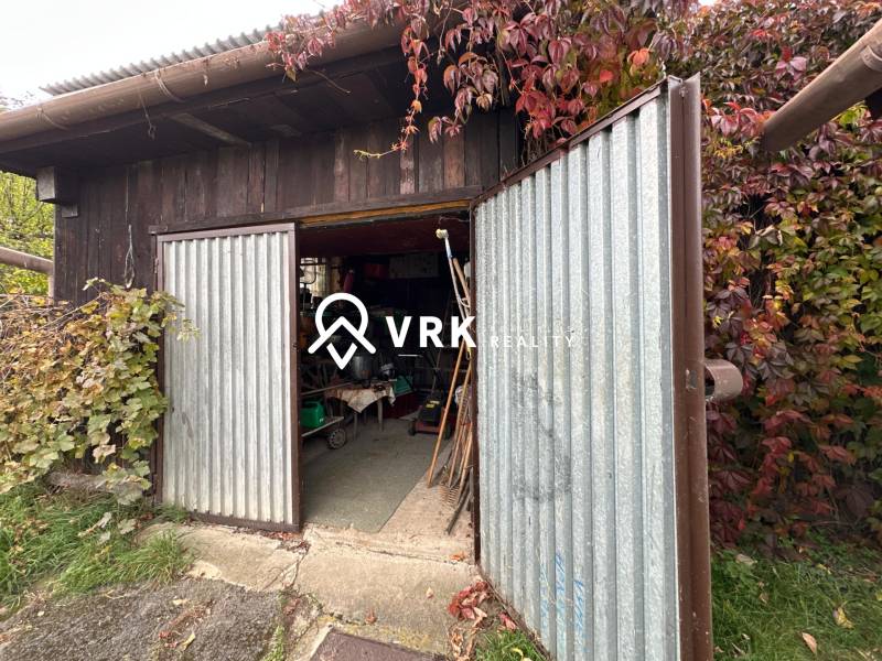 Nyitott fém garázsajtó egy családi háznál Kalša-ban, buja növényzettel körülvéve.