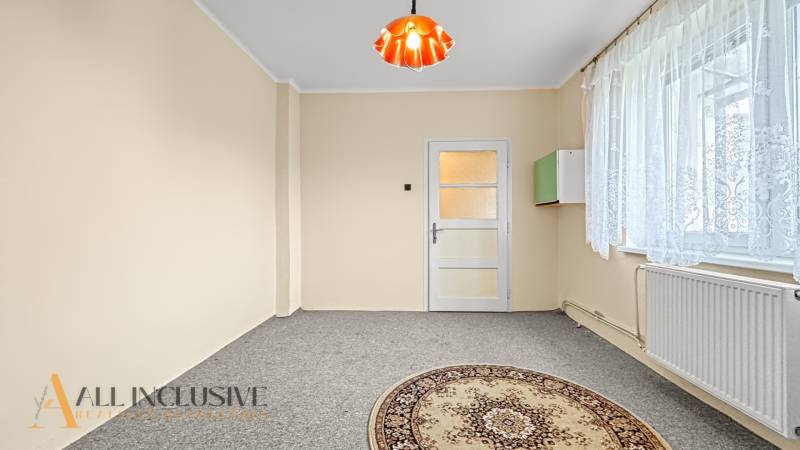 Világos szoba családi házban, szőnyeggel és csipkefüggönyös ablakkal.