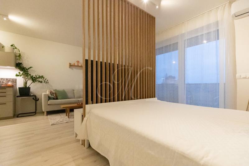 Konyha világos bútorokkal, kanapéval és ággyal egy egyszobás lakásban, fa dekoros padlóval.