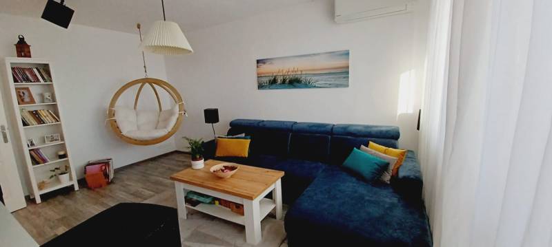 Nappali kék kanapéval, hintaszékkel, könyvespolccal és fa mintázatú padlóval.