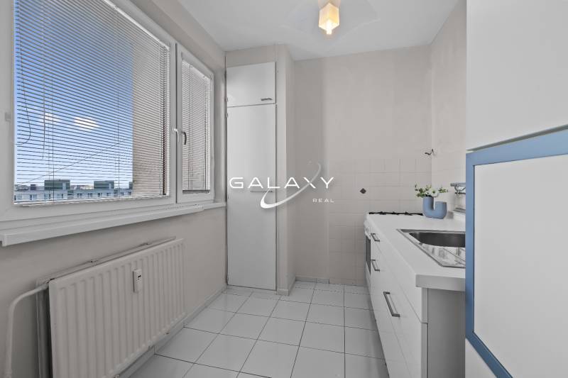 Konyha egy egyszobás lakásban fehér bútorokkal és csempékkel, kilátással a redőnyökön keresztül.