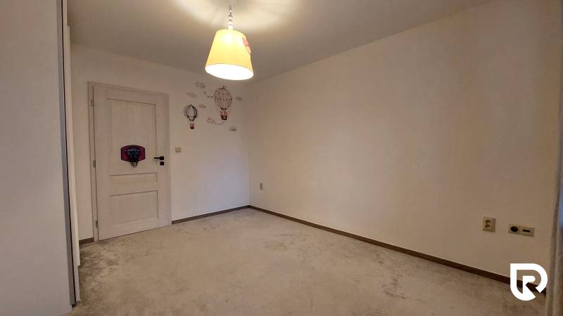 Szoba egy háromszobás lakásban szőnyeggel, világos lámpával és fali dekorációkkal.