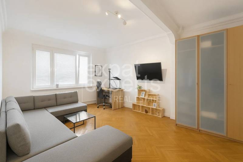 Nappali egy háromszobás lakásban sarokkanapéval, íróasztallal és fa mintázatú padlóval.