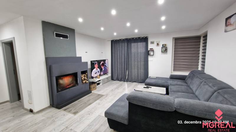 Nappali családi házban kandallóval, szürke kanapéval és televízióval, fa mintázatú padlóval.