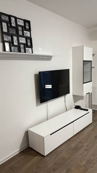 Televízió a falon, fehér komód, képek és fa mintázatú padló az egyszobás lakásban.