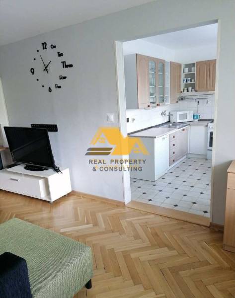 Nappali egy háromszobás lakásban, fa mintázatú padlóval és háttérben konyhával.