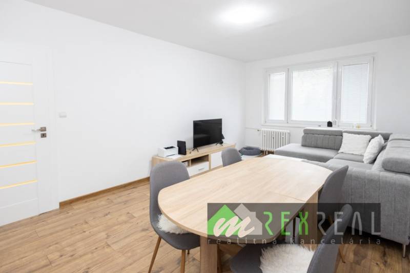 Nappali egy háromszobás lakásban, fa mintázatú padlóval és szürke kanapéval.