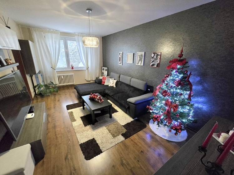 Nappali egy háromszobás lakásban kanapéval, világító fával és fa dekorációjú padlóval.