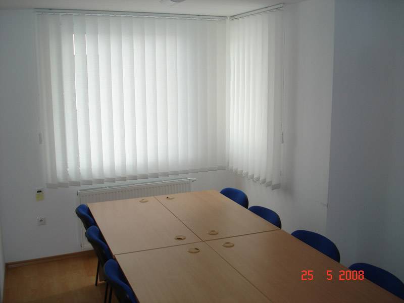 Irodai helyiség tárgyalóasztallal, kék székekkel és függőleges redőnyökkel.