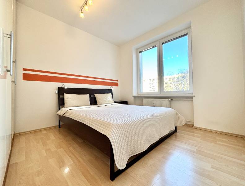 Hálószoba nagy ággyal, fehér falakkal és fa mintázatú padlóval.