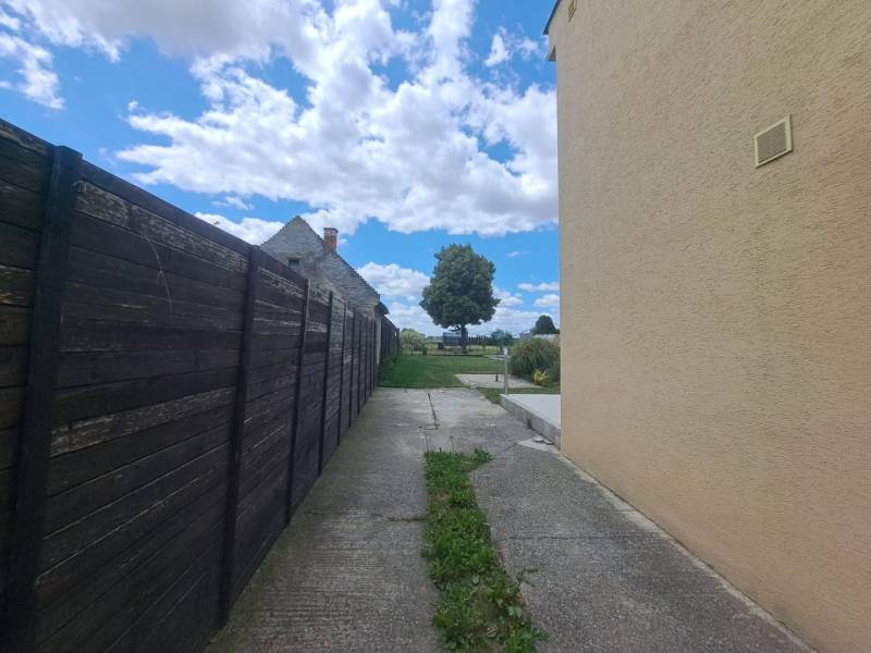 Családi ház Nagycsaládon a fakerítés mellett, betonút, felhők és fa.