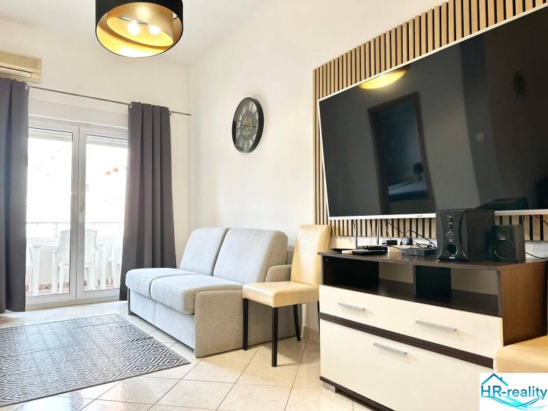 Nappali kanapéval és televízióval egy üdülő apartmanban, világos csempe, erkélyajtók.