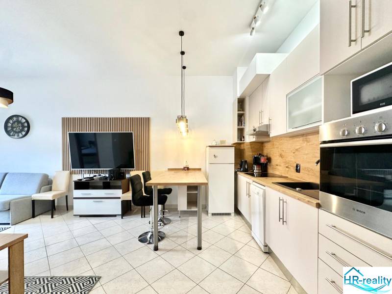 Rekreációs apartman belső tere konyhasarokkal, étkezőasztallal és falra szerelt televízióval.