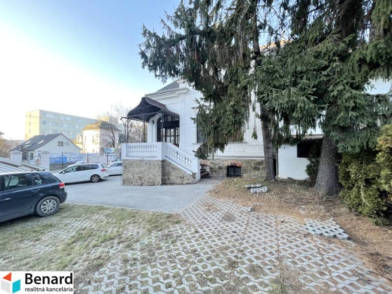Az épület a Vajanského utcában, Eperjesen, a bevezető úttal, autókkal és fenyőfákkal az előtérben.