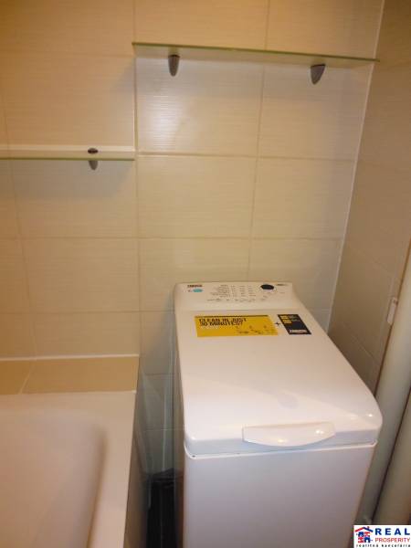 Fürdőszoba egy kétszobás lakásban, mosógéppel a kád mellett és polcokkal.