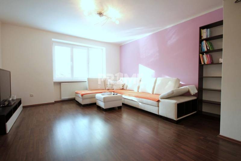 Nappali egy kétszobás lakásban sarokkanapéval, könyvespolccal és fa mintázatú padlóval.