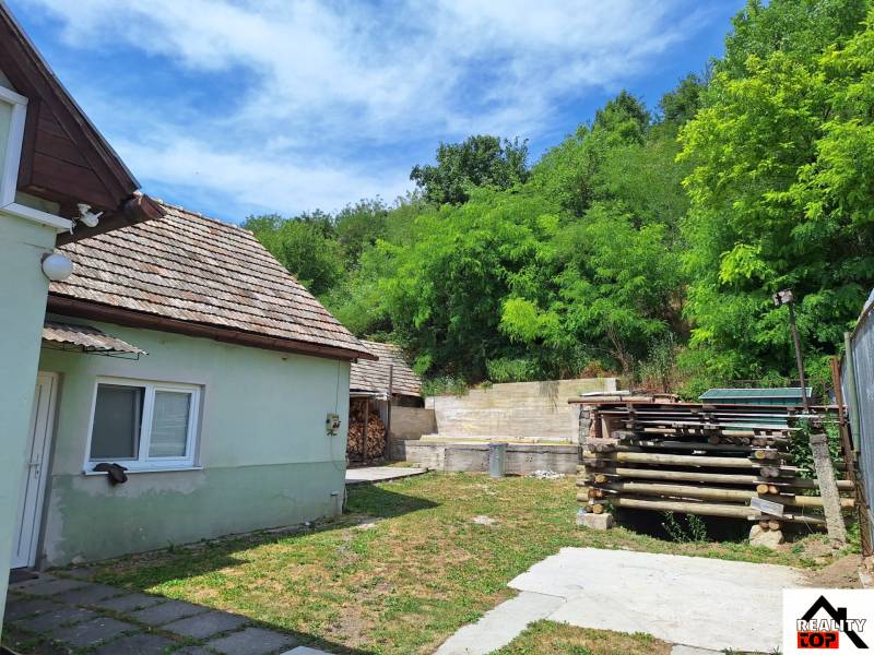 Családi ház Ajnácskőn udvarral, fáskamrával és körülötte zöldellő fákkal.