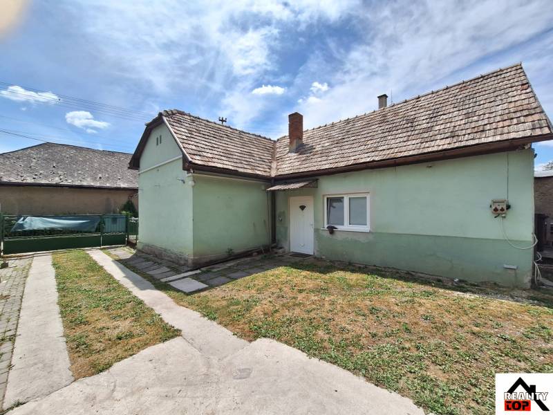 Családi ház Ajnácskőn aszfaltos járdával és zöld gyepes udvarral a kék ég alatt.