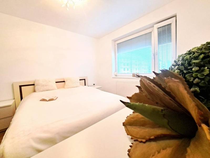 Világos hálószoba nagy ággyal, könyvvel és növénnyel egy kétszobás lakásban.