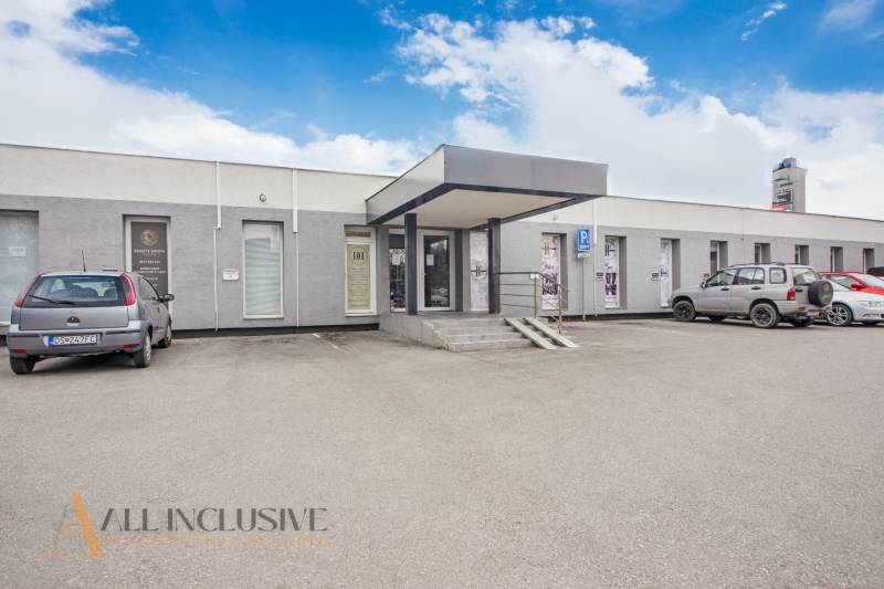 Egyszintes épület üzlethelyiségekkel a Vajanského utcán, parkolóval és autókkal a bejárat előtt.