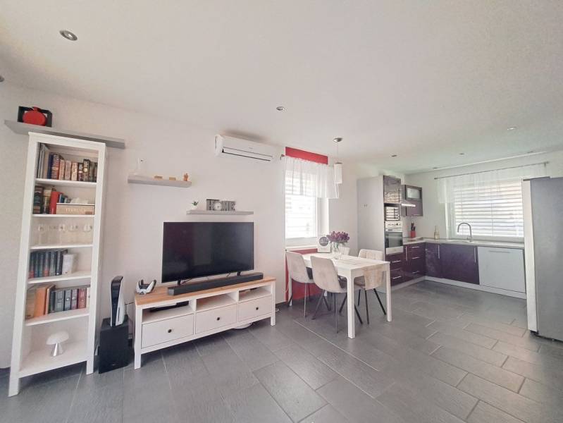 Nappali egy családi házban, televízióval, konyhasarokkal és asztallal, három székkel.