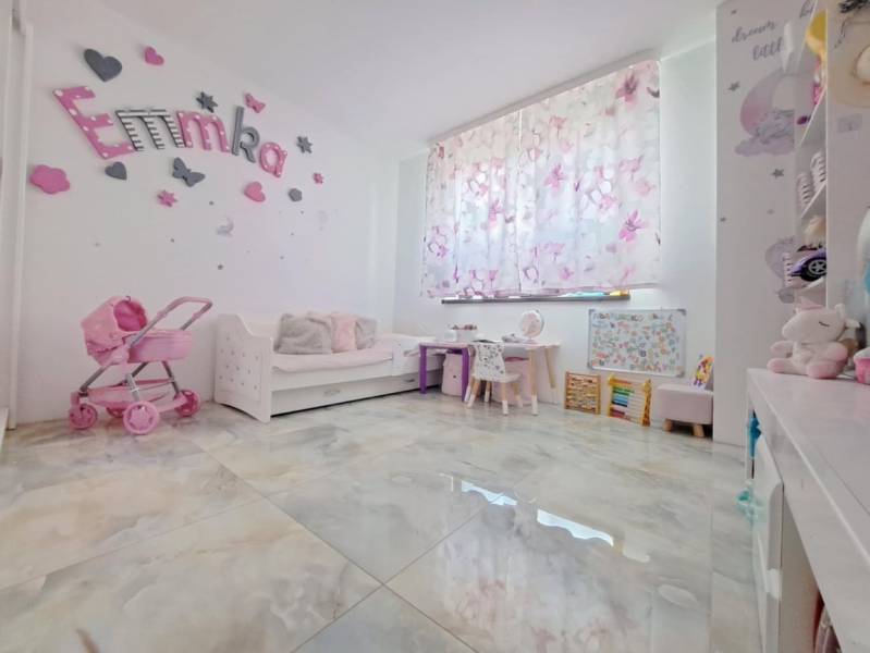 Gyerekszoba egy családi házban dekorációkkal, babakocsival és íróasztalokkal. A padló fa mintázatú.