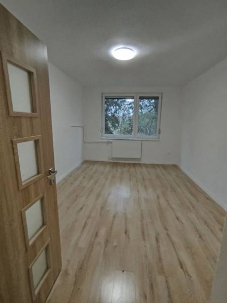 Tágas szoba egy háromszobás lakásban, fa mintás padlóval és nagy ablakkal.