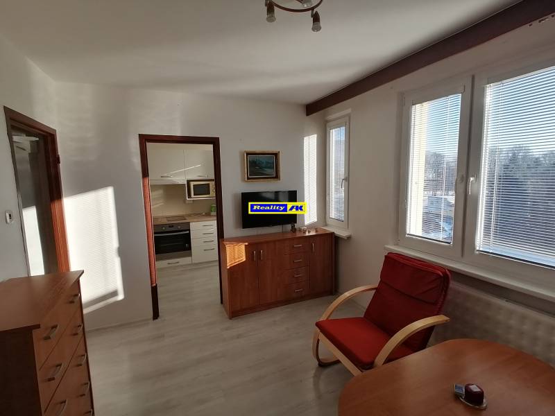 1 szobás lakás fa mintás padlóval, fotellel és konyhába vezető bejárattal.