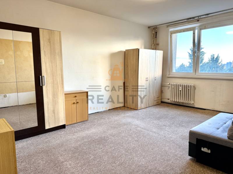 Szoba egy egyszobás lakásban bútorokkal és szőnyeggel, világos falak, nagy ablak.