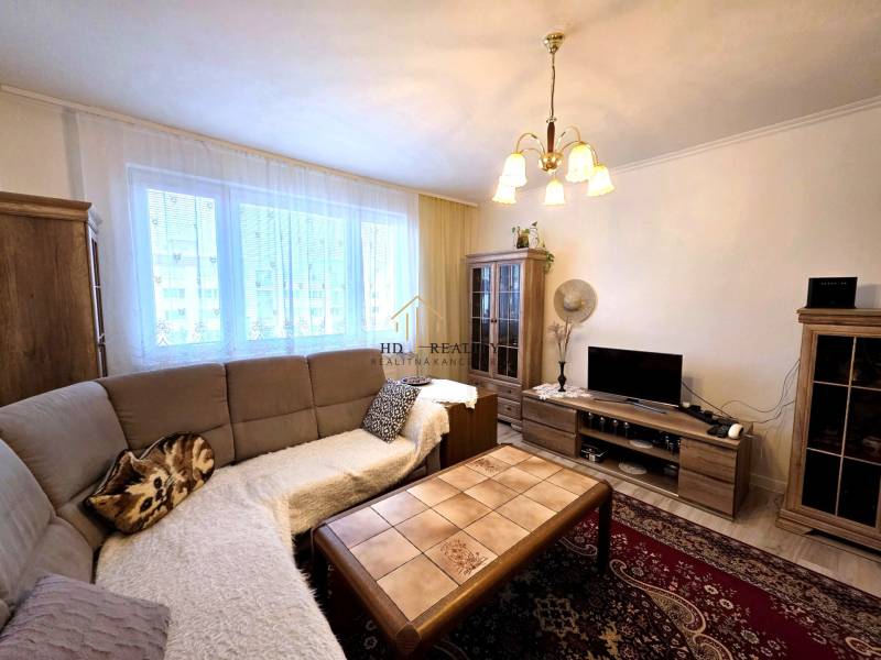 Nappali egy háromszobás lakásban kanapéval, csillárral, fa mintázatú padlóval és szekrényekkel.