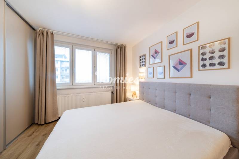 Világos hálószoba fa mintázatú padlóval, ággyal és fali képekkel egy kétszobás lakásban.