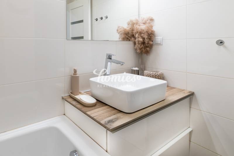 Fürdőszoba egy kétszobás lakásban mosdóval, fa mintázatú padlóval és dekoratív növényi elrendezéssel.