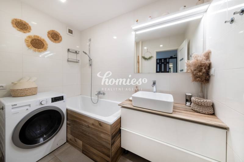 Fürdőszoba egy kétszobás lakásban mosógéppel és káddal, világos csempékkel és dekorációkkal.