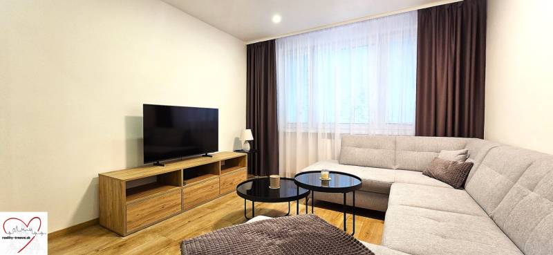 Nappali egy háromszobás lakásban kanapéval, televízióval és dohányzóasztallal, fa dekor padlón.