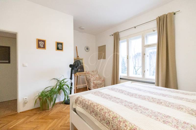 Világos hálószoba fa mintás padlóval, növényekkel és nagy ablakokkal egy kétszobás lakásban.